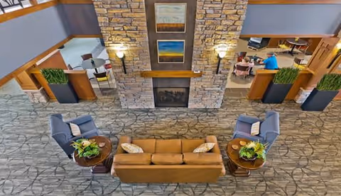A spacious senior living common area with a stone fireplace in the center. The room features a brown sofa and two blue armchairs arranged around two round wooden tables with plants on them. In the background, there are additional seating areas with tables and chairs, and the space is decorated with plants and framed artwork above the fireplace.
