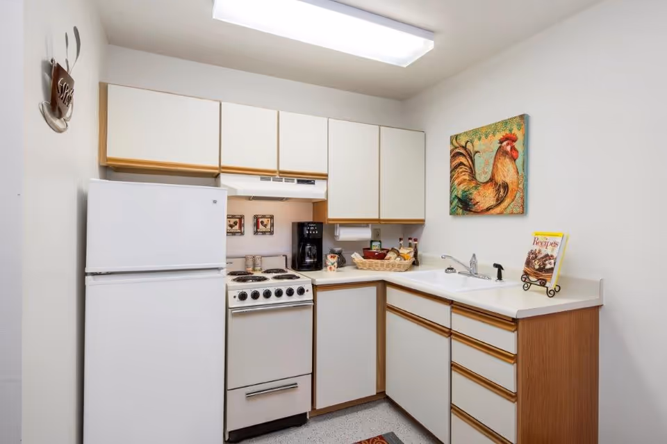 A small kitchen with white cabinets and wooden trim, a white refrigerator, a white stove with four burners, a coffee maker, a double sink, and a countertop with a basket of items and a cookbook on a stand. A colorful rooster painting hangs on the wall above the sink.