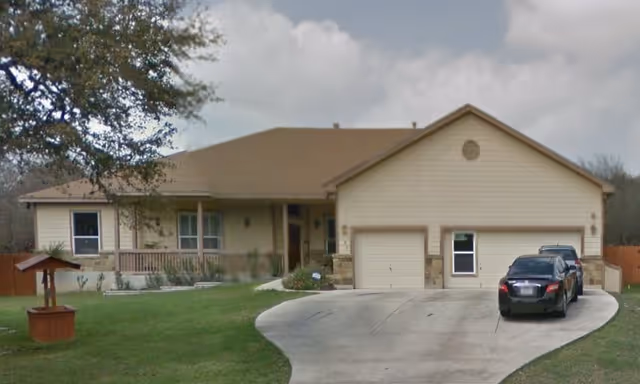 Single-story residential building with a two-car garage, a curved concrete driveway with two parked cars, a small wooden wishing well on the lawn, and a tree on the left side under a partly cloudy sky.