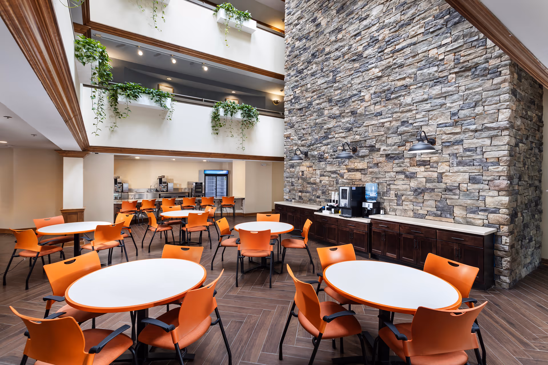 Spacious senior community dining area with round white tables, orange chairs, a stone accent wall, and a beverage station.