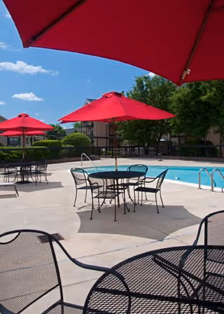 Outdoor pool area with metal tables and chairs under red umbrellas on a sunny day, surrounded by trees and buildings in the background.