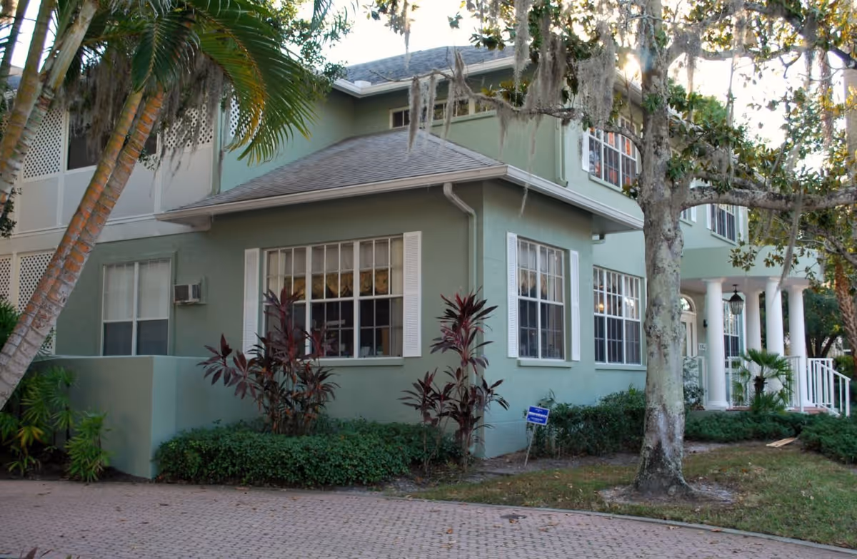 Green two-story building exterior with white-trimmed windows, columns, a front porch, and surrounding trees and landscaping.