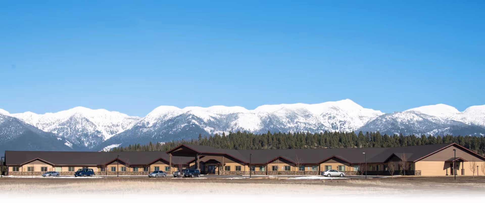 Wide exterior view of Rising Mountains Assisted Living facility with a single-story building featuring a dark roof and beige walls, set against a backdrop of snow-capped mountains and a clear blue sky. Several cars are parked in front of the building.
