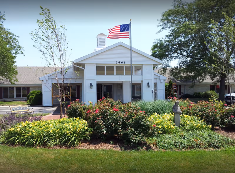 Front view of Franciscan Villa entrance with flag, white building, and blooming garden.