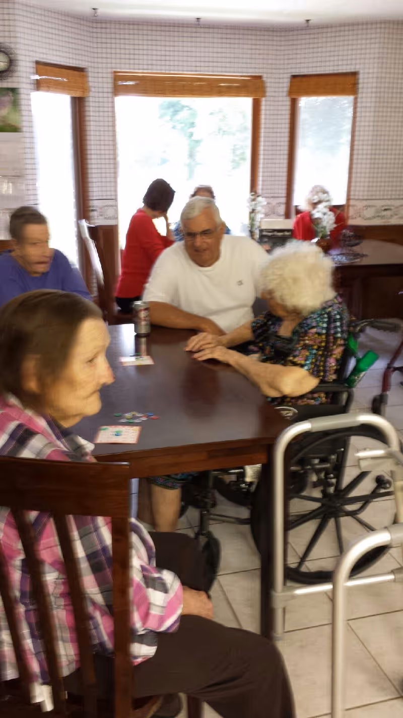 A group of elderly people sitting around a wooden table in a well-lit room with large windows. Some individuals are engaged in conversation, and one person is in a wheelchair. The room has a tiled floor and light-colored walls with a patterned wallpaper border.