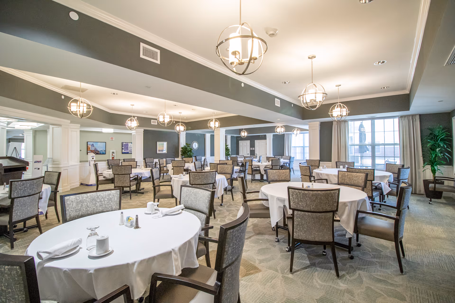 A spacious dining room with multiple round tables covered with white tablecloths, each surrounded by cushioned chairs. The room features modern hanging light fixtures, large windows with curtains, and decorative plants. The walls are painted in a muted green and white color scheme, and framed pictures hang on the walls.