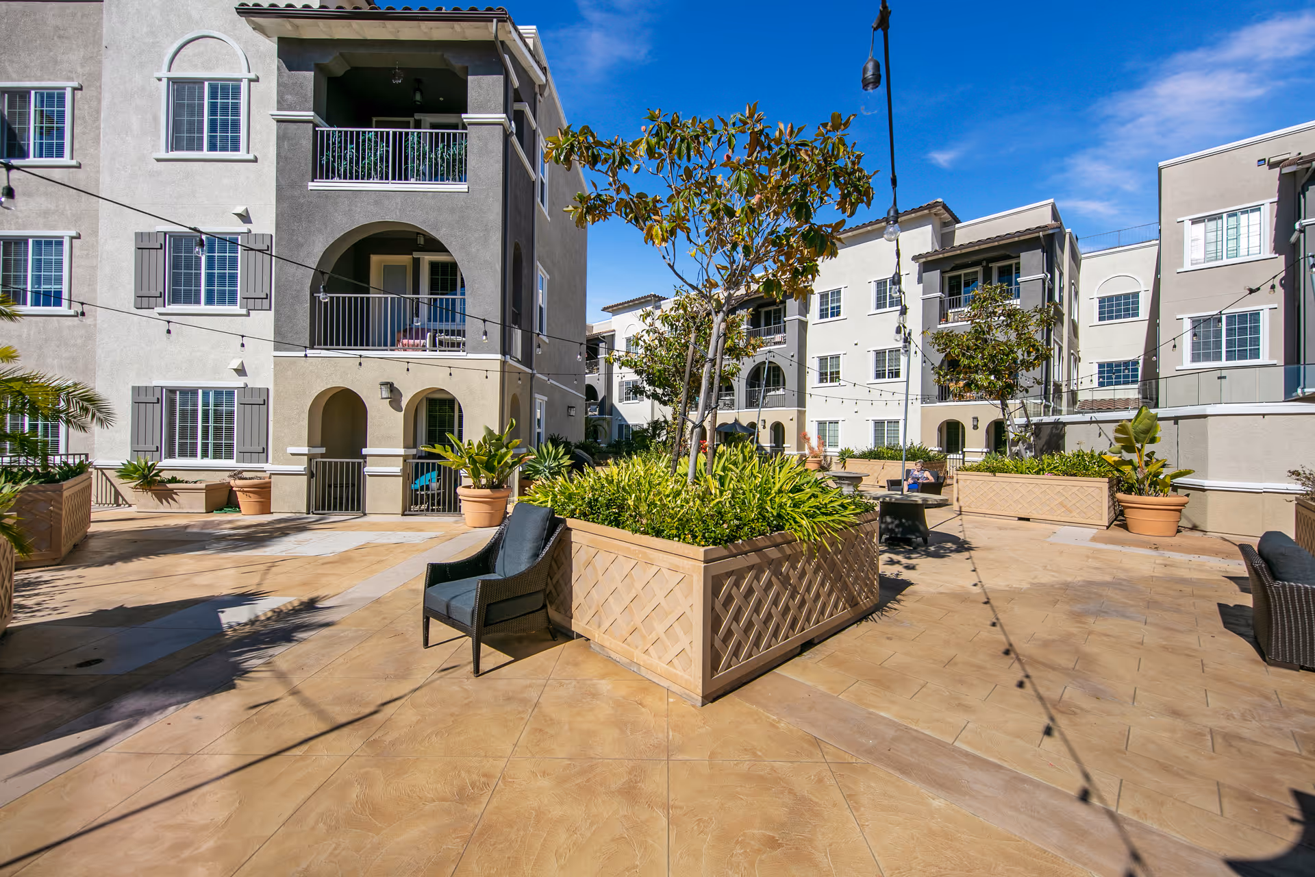 Outdoor courtyard area of a senior living facility with large planters containing trees and greenery, surrounded by multi-story residential buildings with balconies and windows. There are several chairs placed around the courtyard and string lights hanging overhead under a clear blue sky.