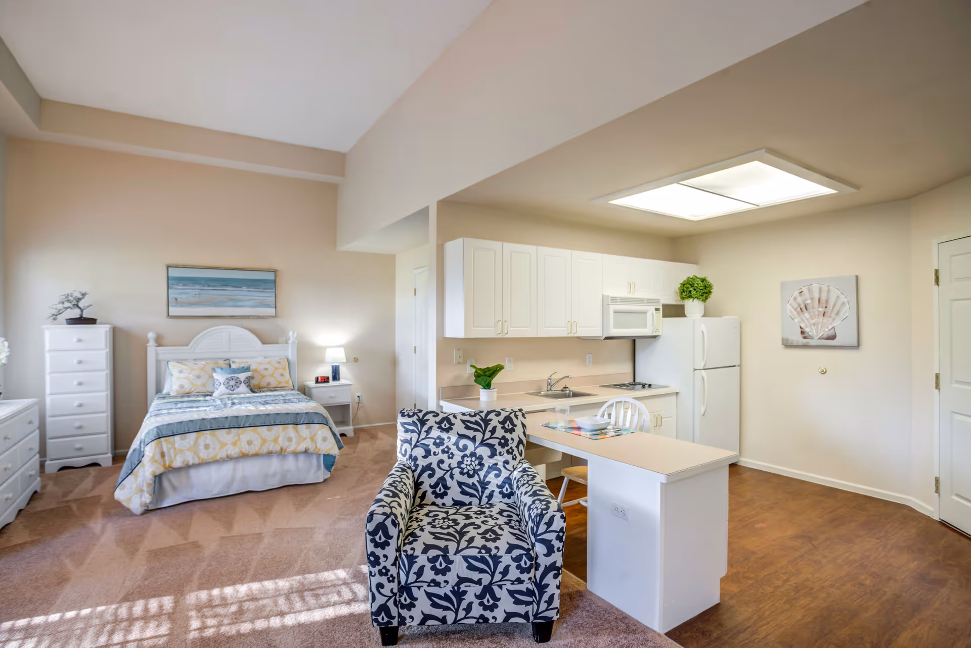 A bright and clean studio apartment featuring a bed with patterned bedding, a white dresser and nightstand with a lamp, a comfortable armchair with a blue and white floral pattern, and a small kitchen area with white cabinets, a refrigerator, microwave, and a countertop with two chairs. The walls are light beige, and there is a painting of a seashell on one wall and a beach scene above the bed.