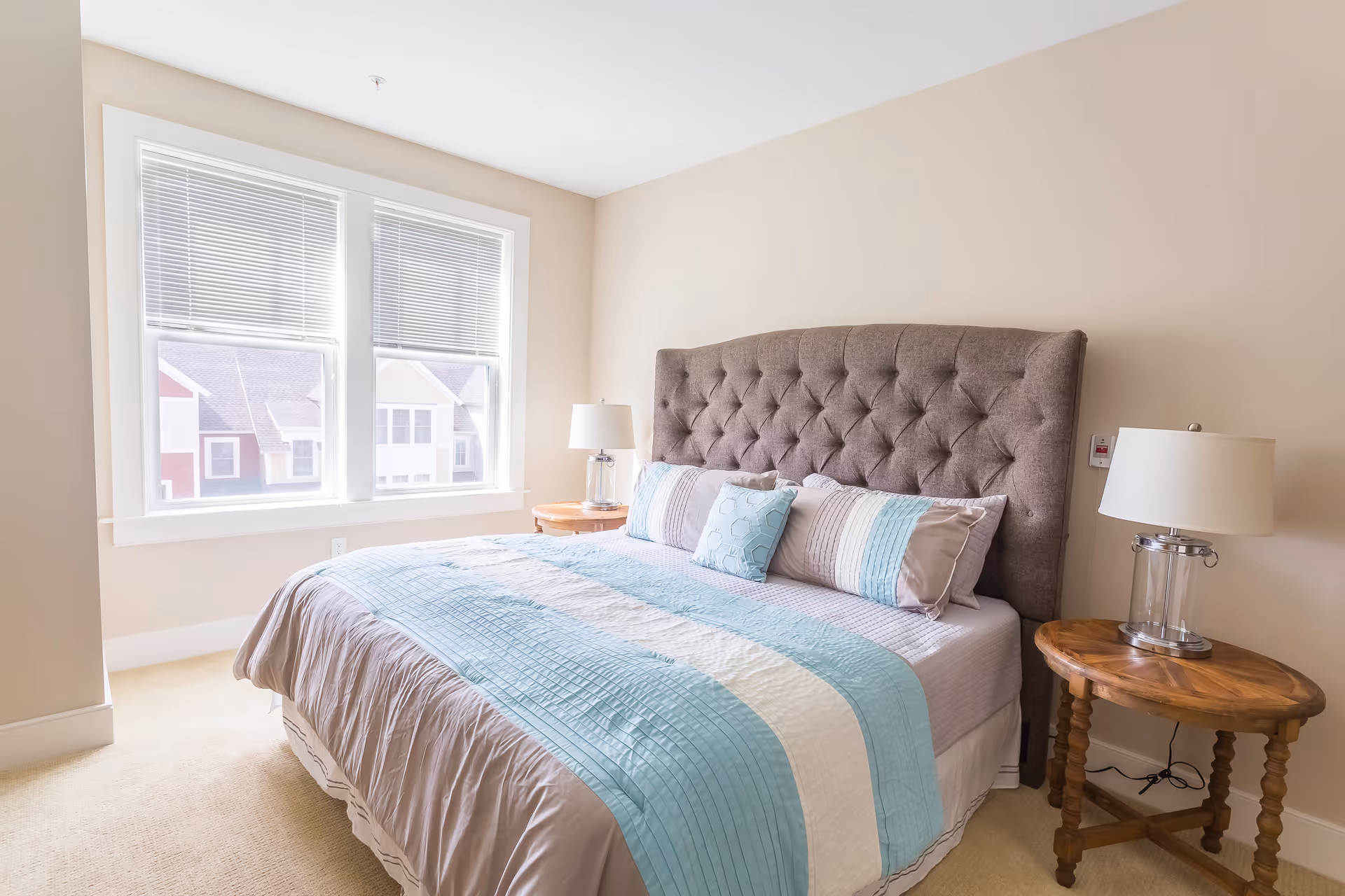A bright bedroom with a large bed featuring a tufted gray headboard and bedding in shades of blue, gray, and white. There are two wooden nightstands on either side of the bed, each with a modern lamp. A window with blinds lets in natural light, and the walls are painted a soft beige color.