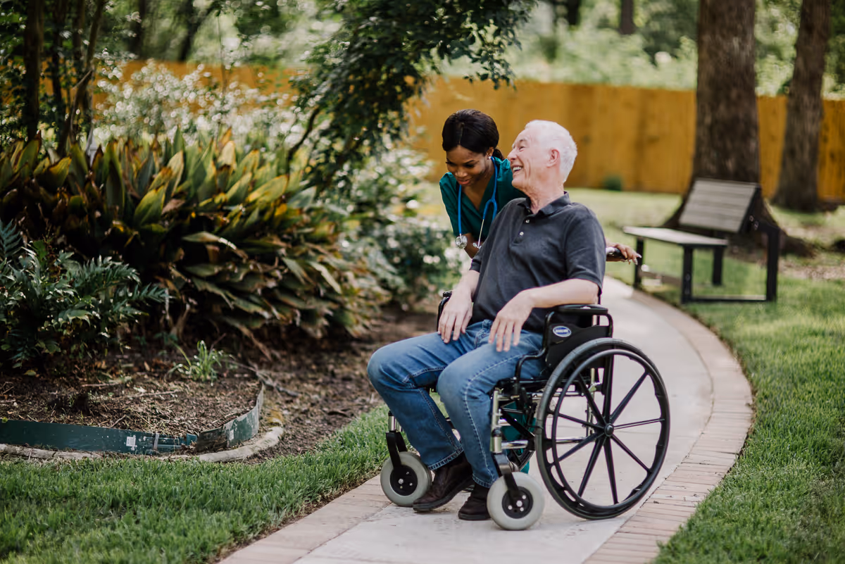 A smiling elderly man in a wheelchair is being assisted by a female healthcare worker wearing scrubs and a stethoscope, outdoors on a paved garden path with greenery and a bench nearby.