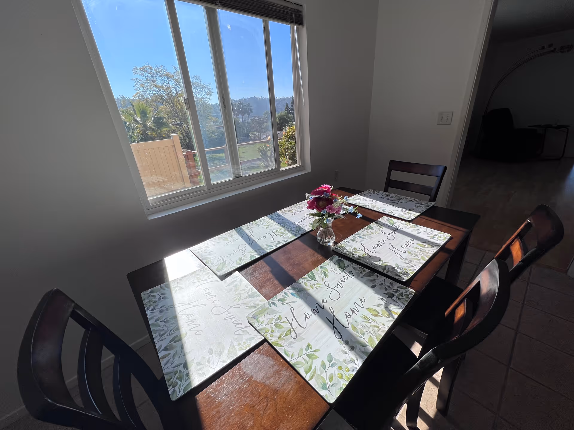 Sunlit dining table with six leafy placemats and a small vase of flowers next to a large window overlooking an outdoor view.