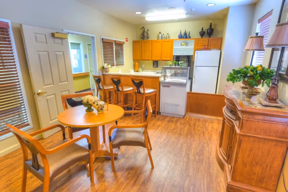 A cozy dining area in a senior living facility featuring a round wooden table with four chairs, a kitchen counter with three bar stools, wooden cabinets, a refrigerator, and decorative items including plants and lamps.