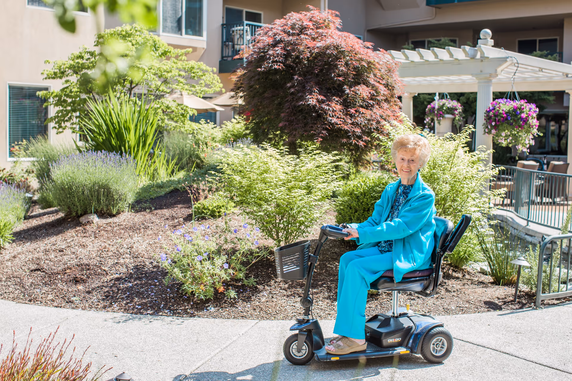 An elderly woman wearing a turquoise outfit is sitting on a mobility scooter on a paved pathway in a garden area with various green plants, flowers, and a pergola in the background.