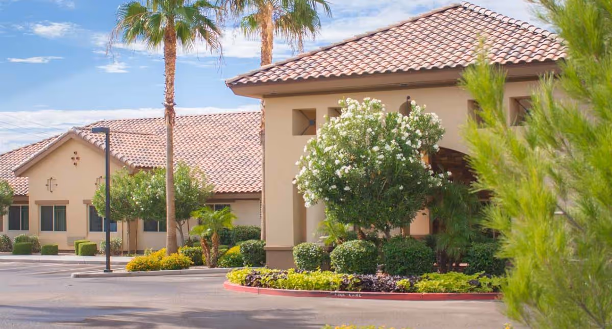 Front entrance of a Mediterranean-style building with a tiled roof, palm trees, and landscaped shrubs and flowers.