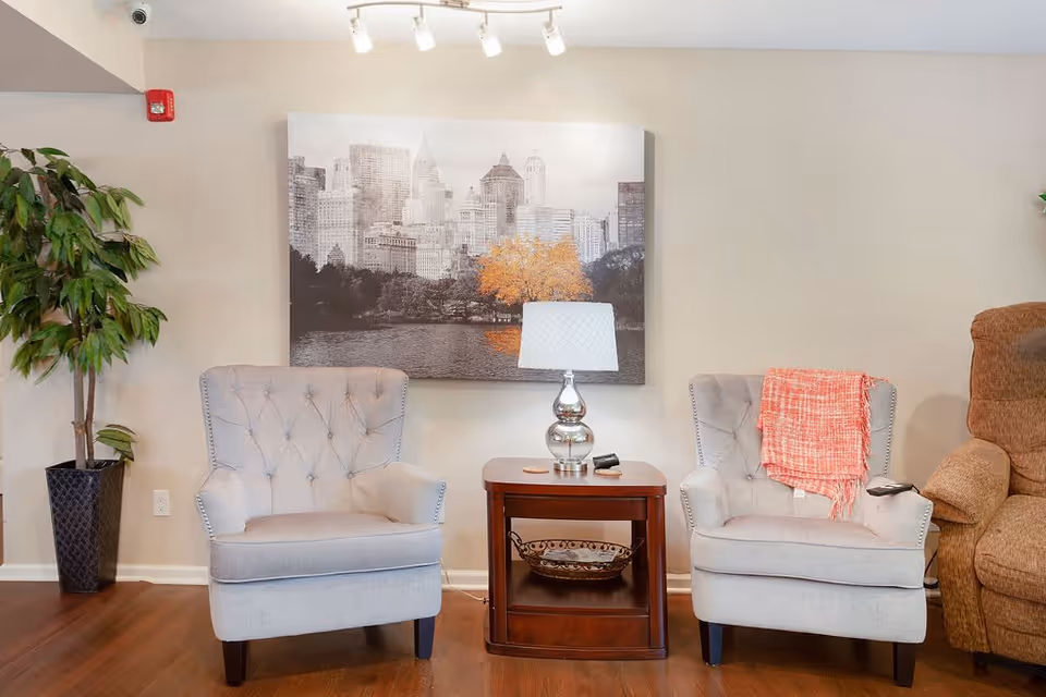 A cozy seating area with two tufted armchairs flanking a wooden side table topped with a lamp beneath a cityscape painting.