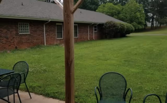 Grassy yard beside a single-story brick building with patio chairs and a wooden post in the foreground.