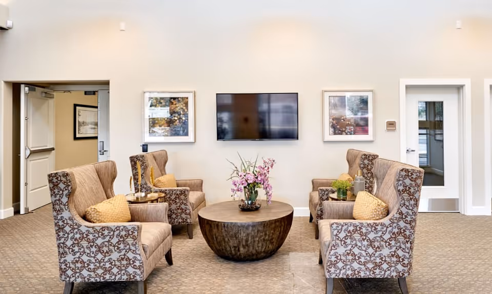 A cozy seating area in a senior living facility with four patterned armchairs arranged around a round wooden coffee table with a flower arrangement. A flat-screen TV is mounted on the beige wall, flanked by two framed pictures. There are two doors visible, one open and one closed, and the floor is carpeted.