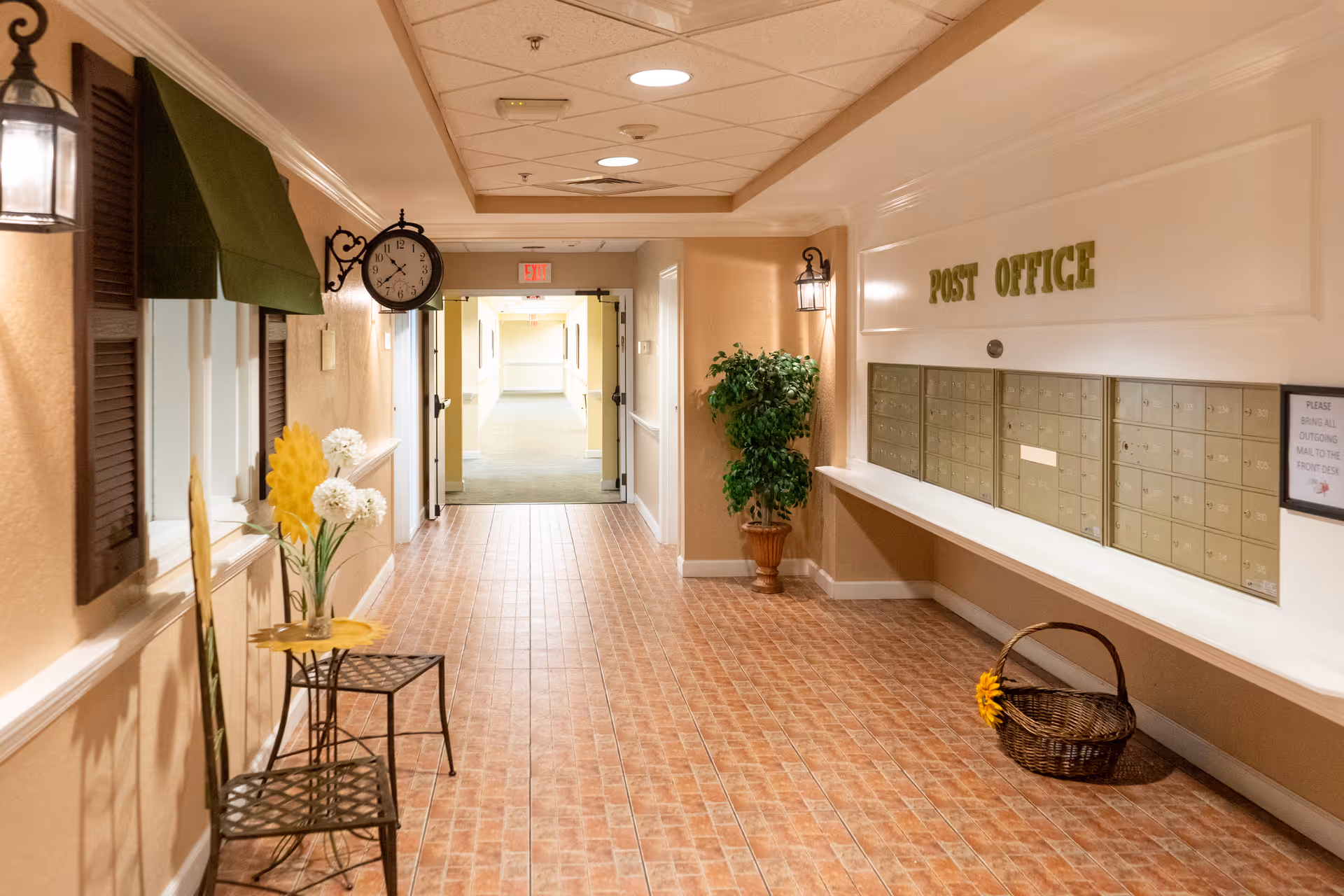 A well-lit interior hallway with a wall of mailboxes labeled 'POST OFFICE', decorative seating, plants, and a hanging clock.