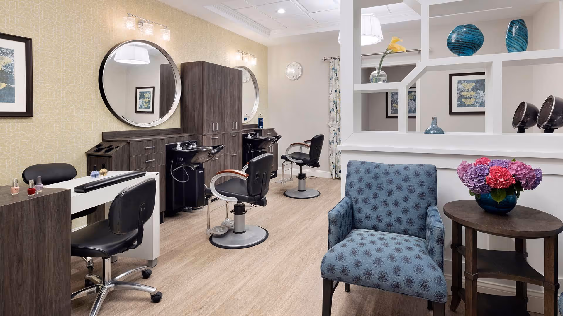Interior view of a salon area in a senior living facility featuring two black salon chairs in front of wash basins with large round mirrors above. There is a manicure station with a black chair and nail polish bottles on the left. A blue patterned armchair and a small round wooden table with a vase of colorful flowers are in the foreground. The room has light wood flooring, light-colored walls, and decorative framed artwork.