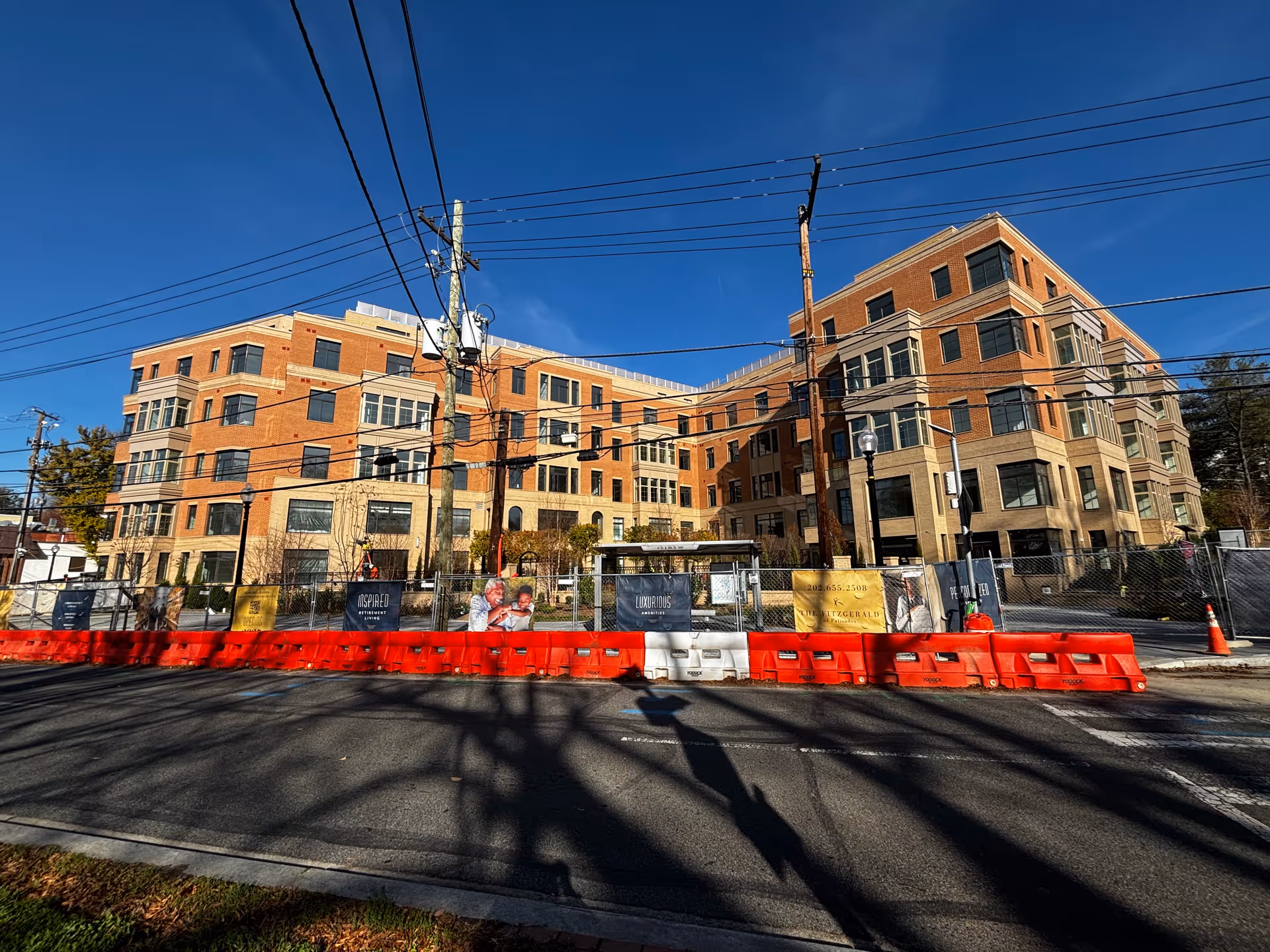Exterior view of a large multi-story brick building under construction or renovation, with orange and white barricades and a chain-link fence in front. Several signs are attached to the fence, including one that reads 'The Fitzgerald of Pallisades'. The sky is clear and blue with visible power lines crossing the scene.