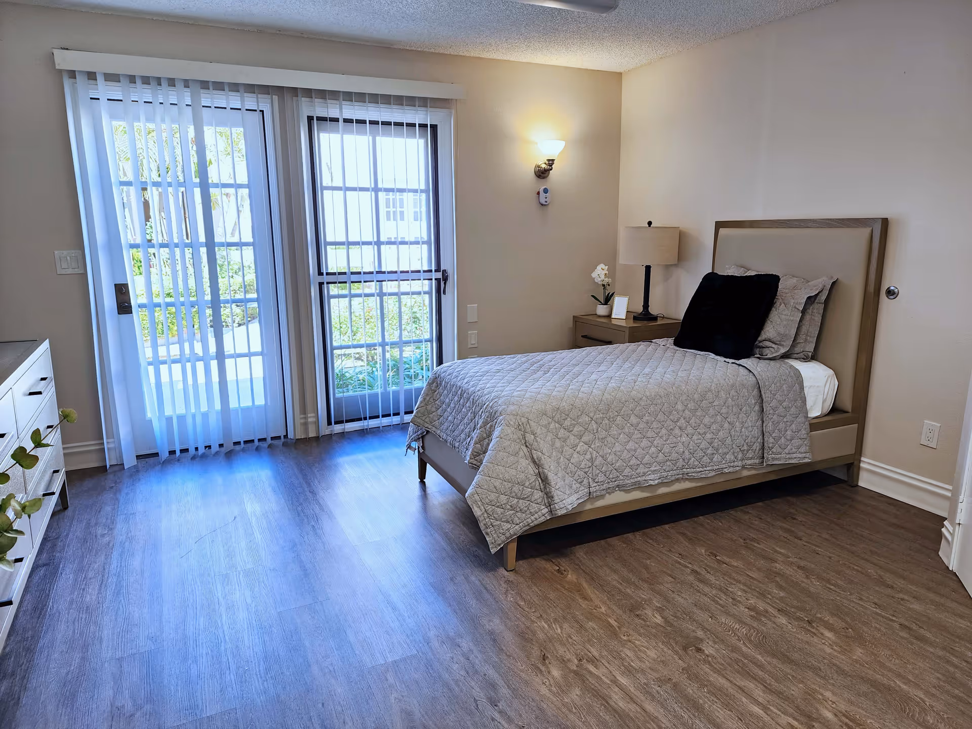 A bright and clean bedroom with a single bed covered in a gray quilt and black pillow. There is a nightstand with a lamp and a small plant next to the bed. The room has wood flooring and large glass doors with vertical blinds that open to an outdoor area with greenery.