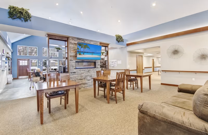 Open common area with wooden tables and chairs, a stone fireplace with wall-mounted TV, and a couch in a senior living facility.