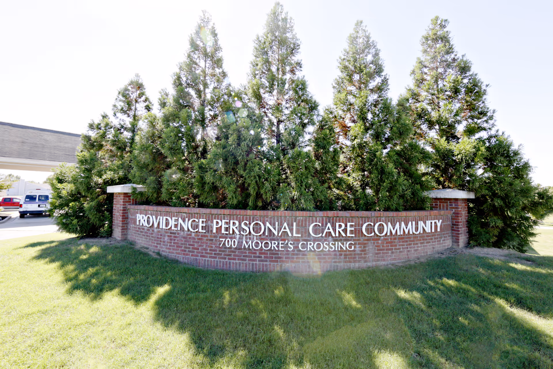 Brick entrance sign reading 'Providence Personal Care Community 700 Moore's Crossing' in front of tall evergreen trees and a grassy lawn.
