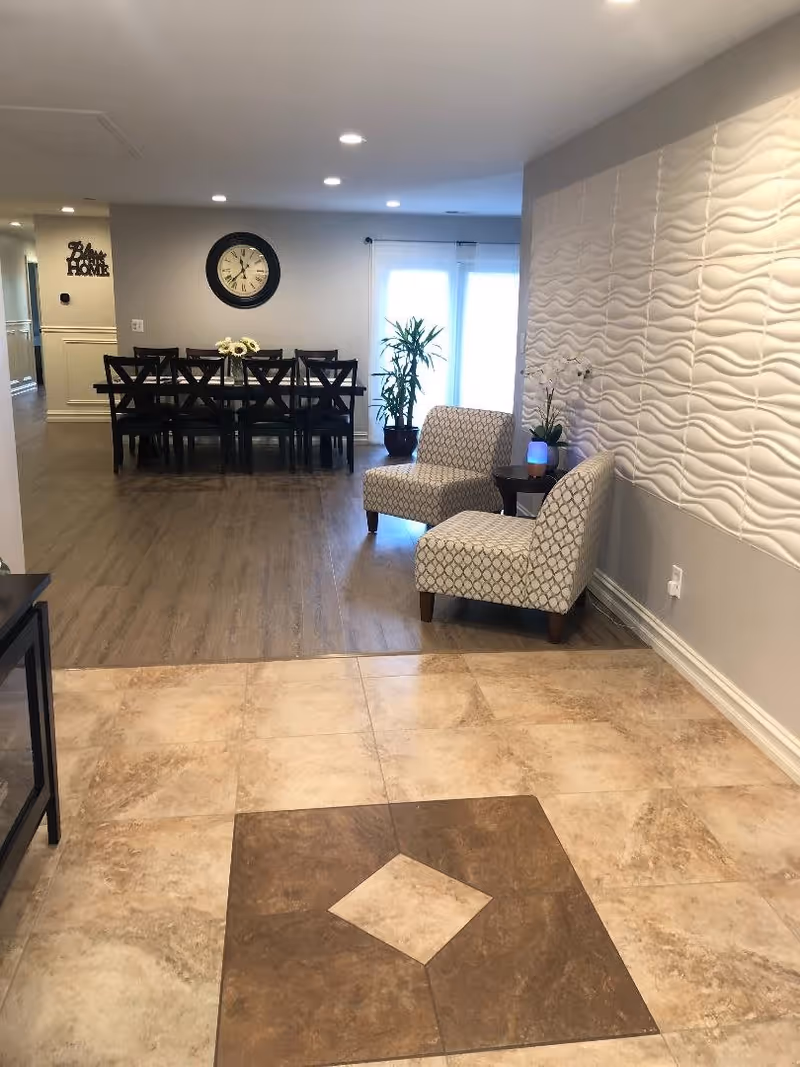 Interior view of a senior living facility showing a tiled floor area leading to a wooden floor dining area with a table and eight chairs. Two patterned armchairs are placed near a textured white wall with a small round table holding a plant and a glowing light. A large clock hangs on the wall above the dining table, and a potted plant is near glass doors letting in natural light.