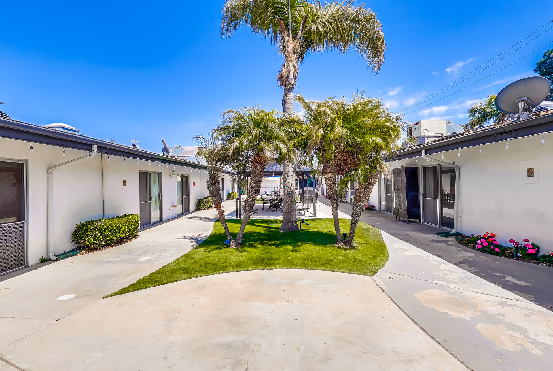 Outdoor courtyard area at Beachside Post Acute featuring a small grassy island with several palm trees in the center, surrounded by a concrete walkway. Single-story white buildings with sliding glass doors and small plants line both sides of the courtyard. The sky is clear and blue with a few clouds.