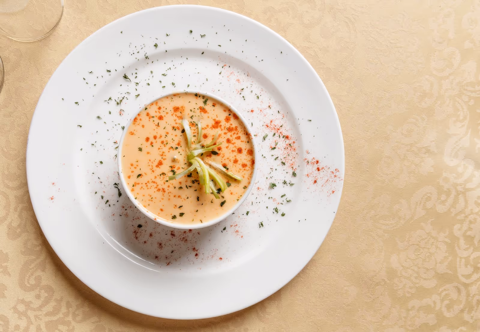 A bowl of creamy soup garnished with chopped herbs and thinly sliced green onions, served on a white plate with sprinkled herbs and paprika on the rim, placed on a beige patterned tablecloth.