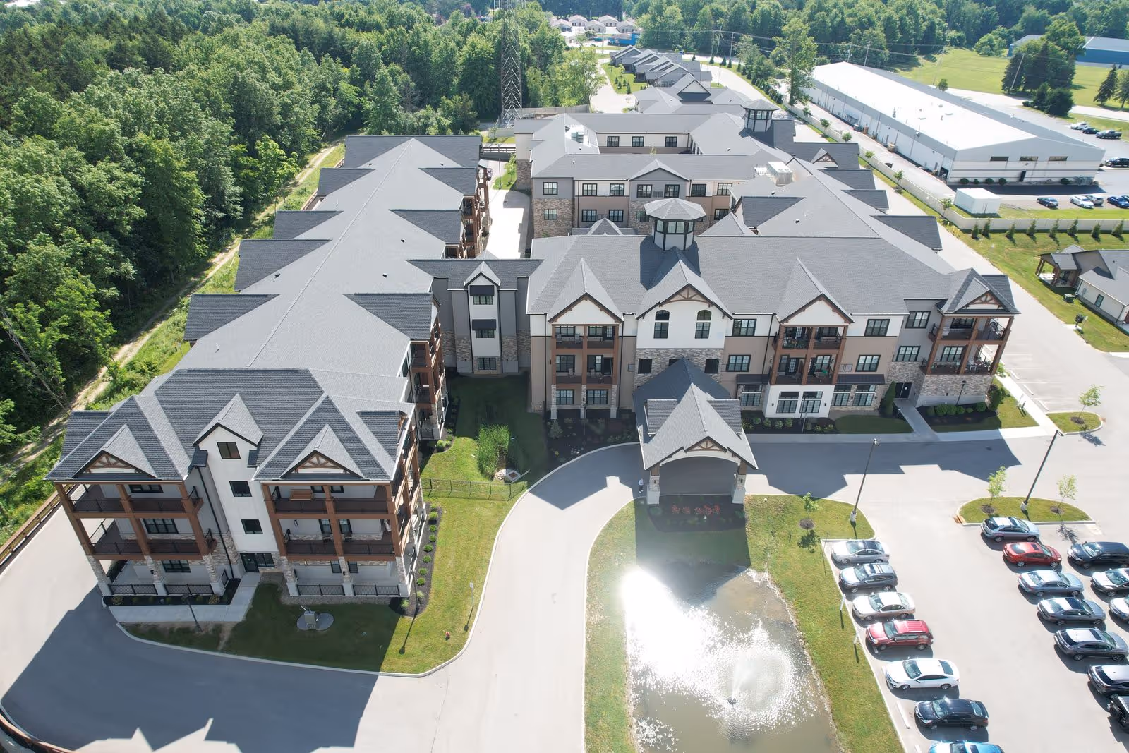Aerial view of a large senior living facility with multiple connected buildings featuring pitched roofs and balconies. The facility is surrounded by greenery and trees on one side, with a parking lot filled with cars on the other. There is a small pond with a fountain near the entrance driveway.
