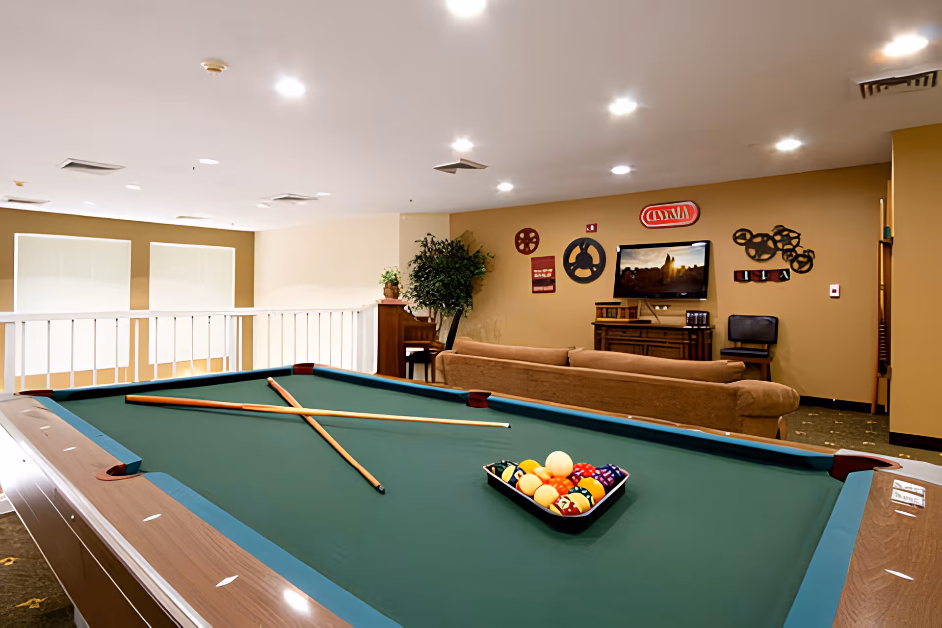 A recreational room with a pool table in the foreground, two pool cues crossed on the table, and a rack of billiard balls. In the background, there is a brown couch facing a wall-mounted TV, with decorative film reels and cinema-themed wall art. A potted plant and a wooden cabinet are also visible.
