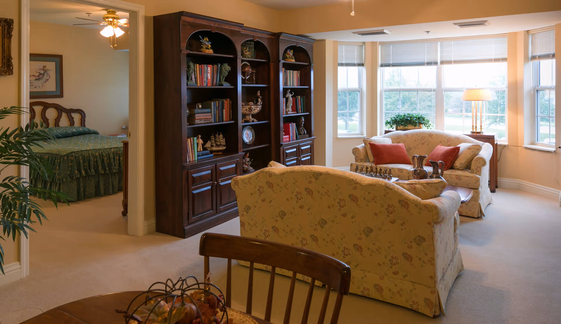 A cozy living room area with floral patterned sofas, a wooden coffee table with decorative items, a large dark wood bookshelf filled with books and ornaments, and large windows letting in natural light. To the left, an open doorway reveals a bedroom with a green bedspread and a ceiling fan.