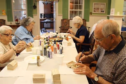Four elderly individuals seated around a table engaged in painting small wooden birdhouses with various colors of paint bottles on the table in a well-lit room.