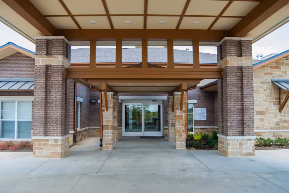 Entrance of Cedar Bluff Assisted Living & Memory Care facility showing a covered drop-off area with brick and stone pillars, glass double doors, and a beige and brown color scheme.