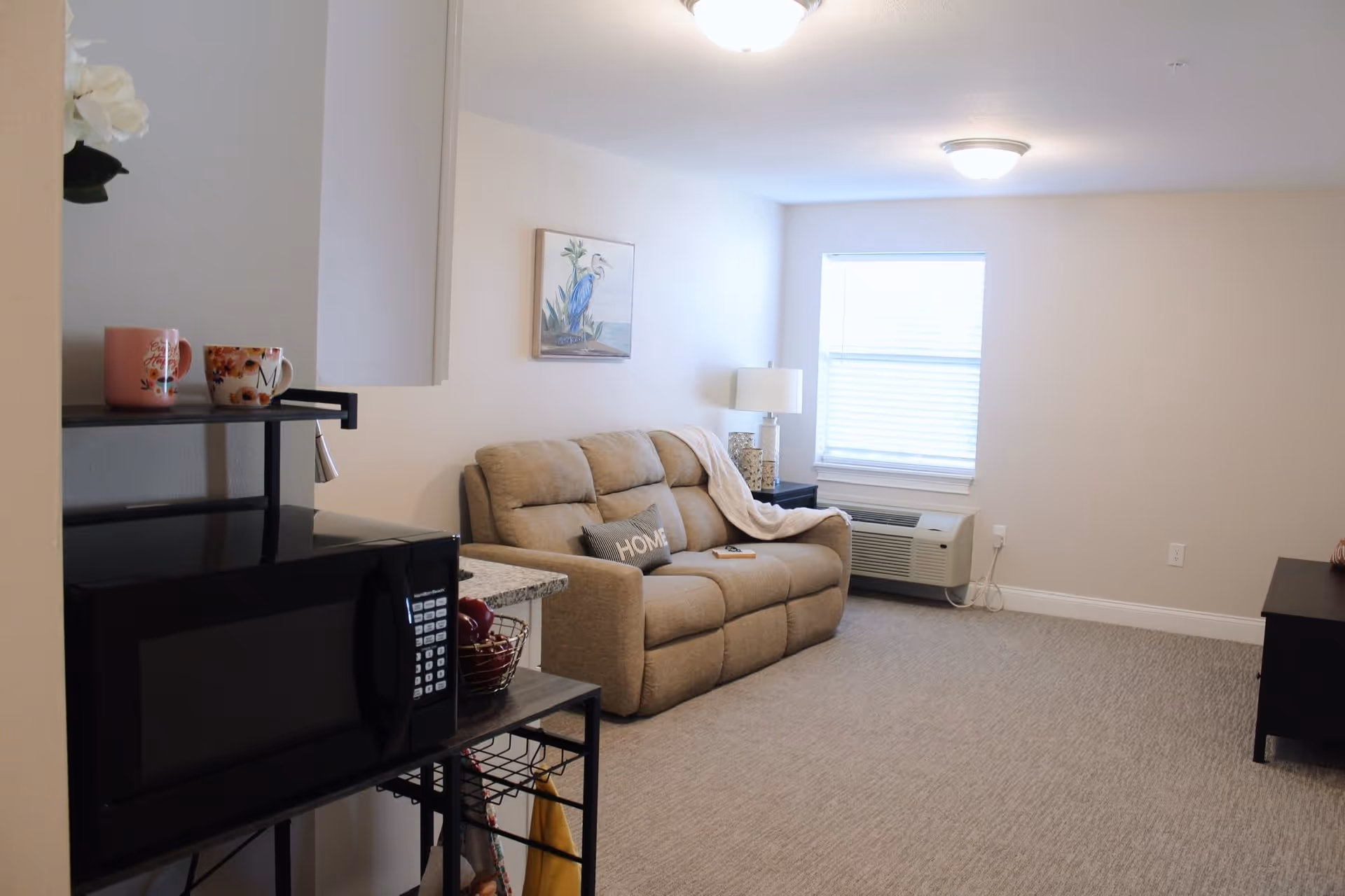A cozy living room with a beige recliner sofa adorned with a 'HOME' pillow and a light throw blanket. A window with blinds lets in natural light above a wall-mounted air conditioning unit. A side table with a lamp and decorative items sits next to the sofa. In the foreground, a black microwave is placed on a metal shelf with two colorful mugs on top. The room has light-colored walls and carpeted flooring.