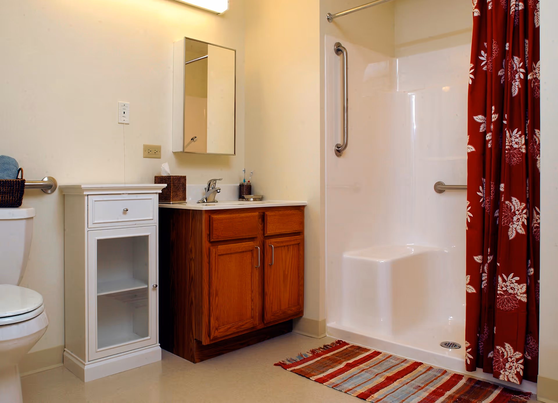A bathroom featuring a white toilet, a white cabinet with a glass door, a wooden vanity with a sink and faucet, a wall-mounted mirror above the sink, and a walk-in shower with a built-in seat and a red floral shower curtain. There is a striped rug on the floor in front of the shower.