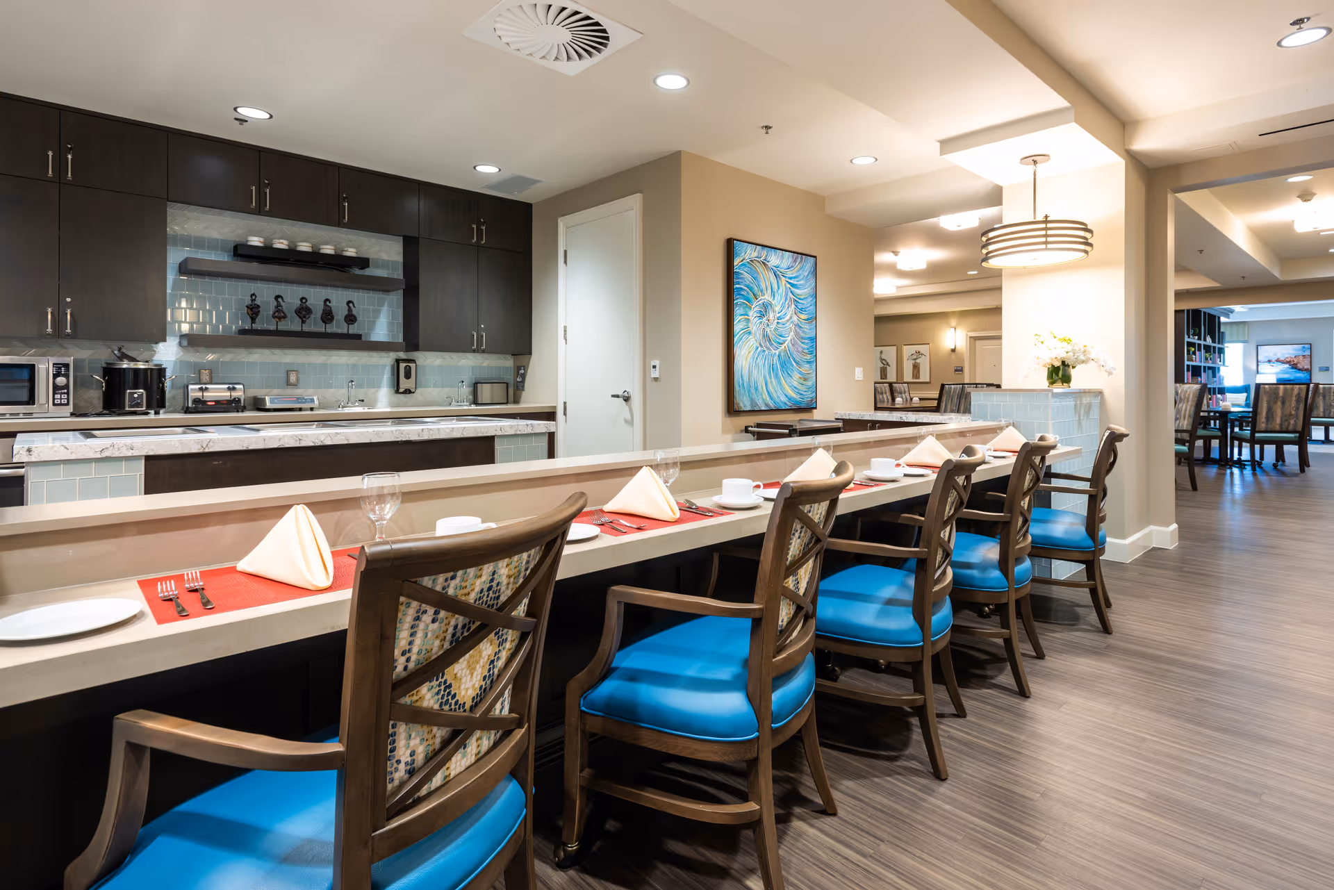 A modern dining area in a senior living facility featuring a long counter with six wooden chairs with blue cushions. The counter is set with red placemats, white plates, folded napkins, cups, and glasses. Behind the counter is a kitchen area with dark cabinets, a microwave, toaster, and other appliances. The room has wood flooring, neutral-colored walls, and a colorful abstract painting on the wall. Additional dining tables and chairs are visible in the background.