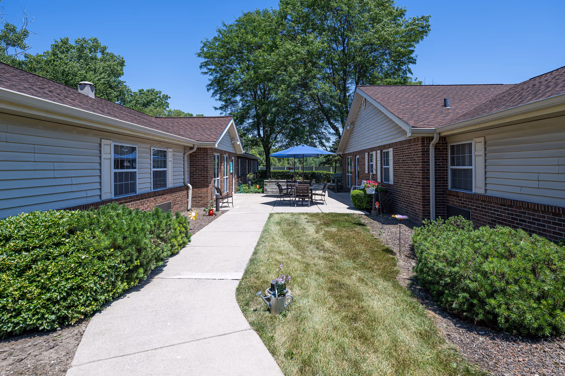 Outdoor courtyard area between two single-story brick and siding buildings with a concrete walkway, green bushes, and a grassy area. At the end of the walkway, there is a patio with a table, chairs, and a blue umbrella under a clear blue sky.