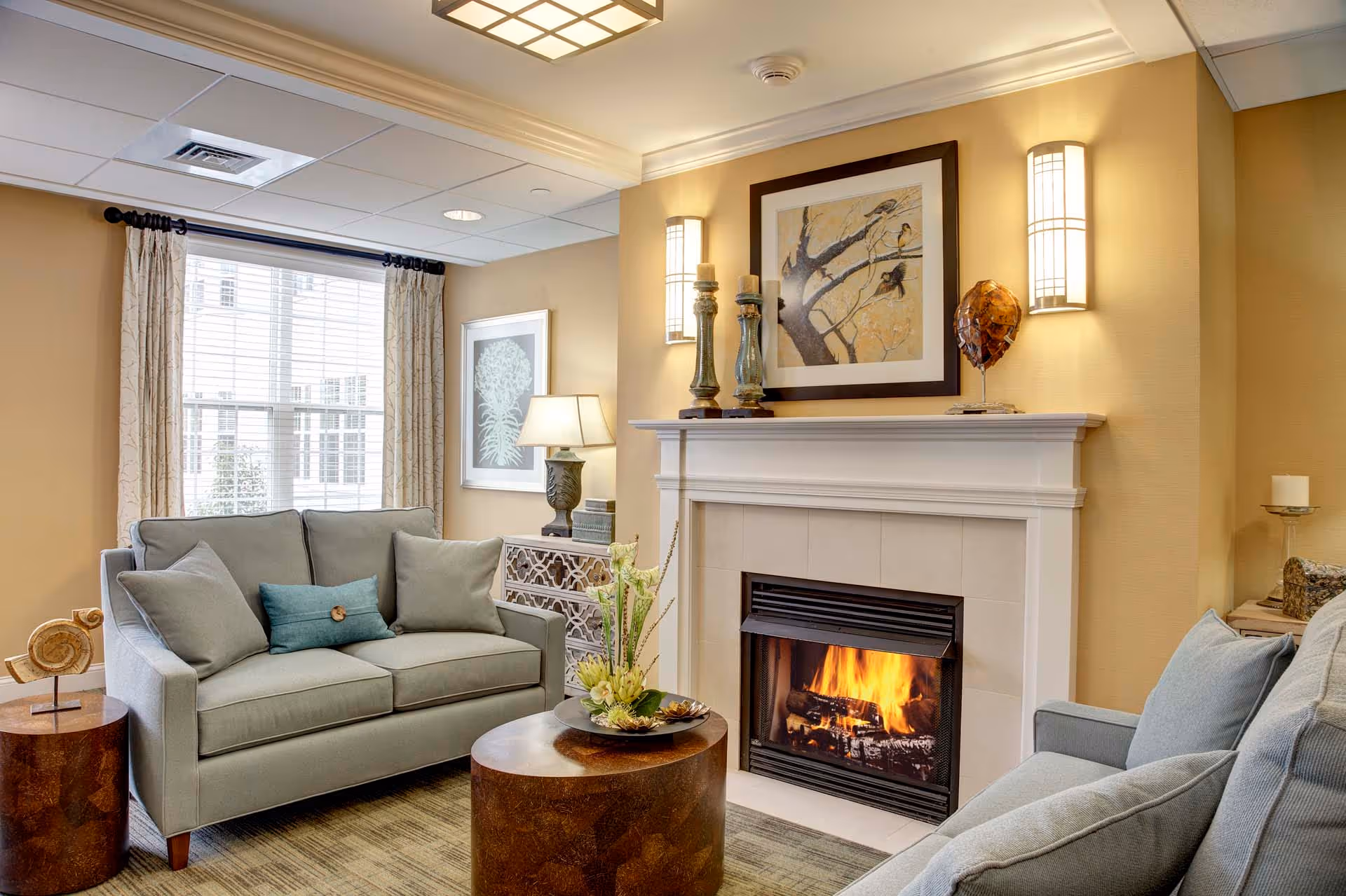 A cozy living room with two light gray sofas facing each other, a round wooden coffee table with a floral arrangement, a lit fireplace with a white mantel, wall sconces, framed artwork above the fireplace, a window with curtains, and a side table with a lamp and decorative items.