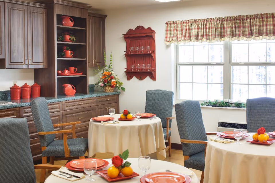 A dining room with round tables covered in beige tablecloths, each set with red plates, glasses, and napkins. The chairs around the tables have wooden arms and blue upholstery. In the background, there are wooden cabinets with red kitchenware and a red wall-mounted shelf holding glasses. A window with a plaid valance lets in natural light, and a floral arrangement is placed on the countertop.