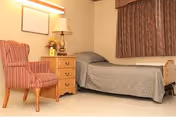 A simple bedroom with a single bed covered with a gray quilt, a wooden nightstand with a lamp and flowers, a striped upholstered chair, and a window with patterned curtains.