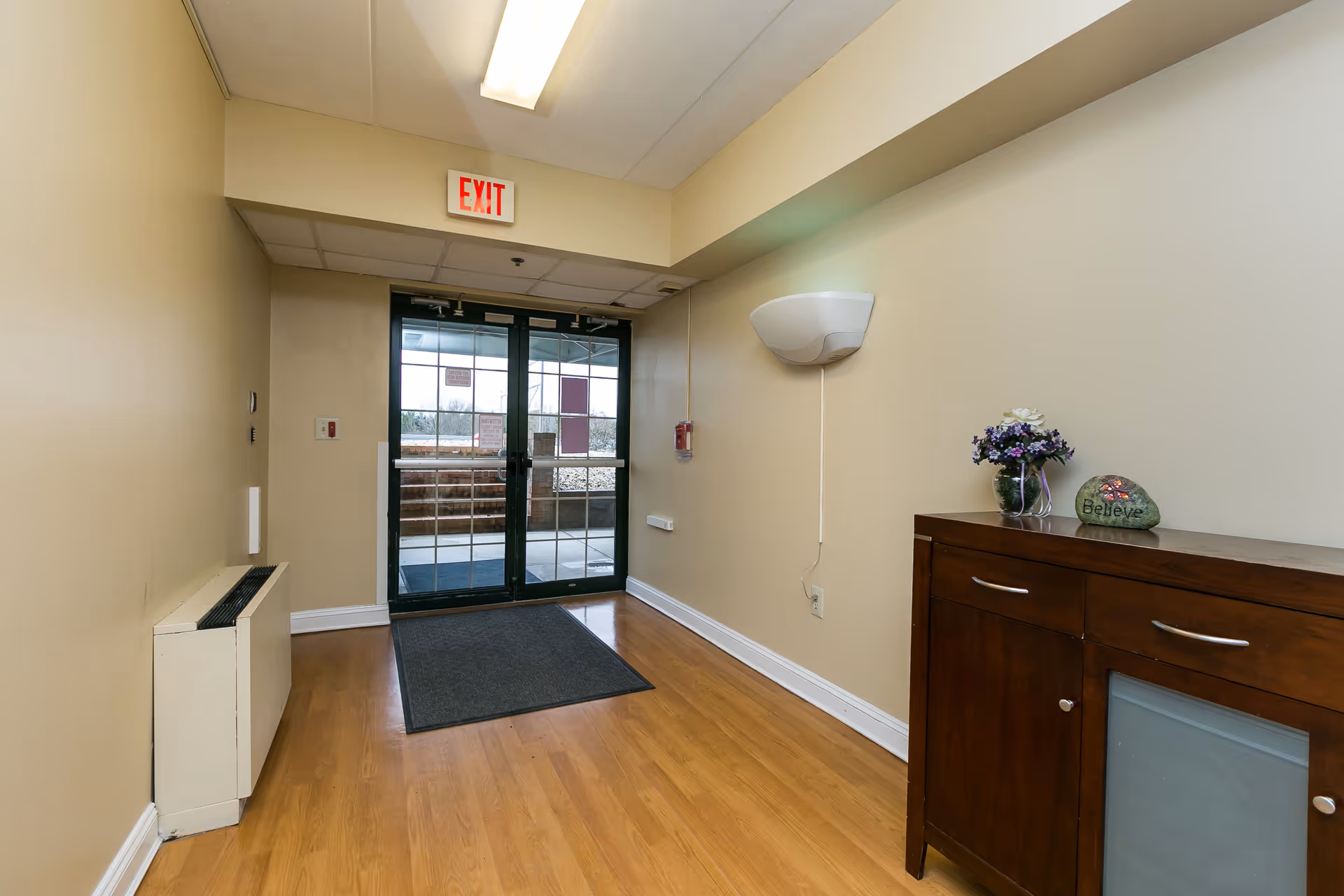 Interior hallway with light wood flooring leading to glass double doors with an exit sign above. On the right side, there is a wooden cabinet with a small vase of purple flowers and a decorative rock with the word 'Believe'. The walls are painted beige and there is a wall-mounted light fixture and a fire alarm on the right wall.