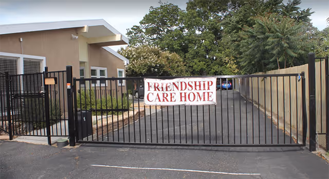 Entrance gate to Friendship Care Home with a black metal fence and a white banner displaying the facility name. A beige building is visible on the left side behind the gate, with trees and a driveway extending beyond the gate.