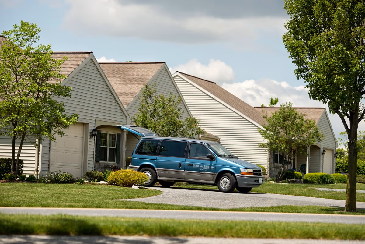 A blue van with the Garden Spot Village logo parked in the driveway of a beige residential building with a garage. The building is surrounded by green grass, bushes, and trees under a partly cloudy sky.