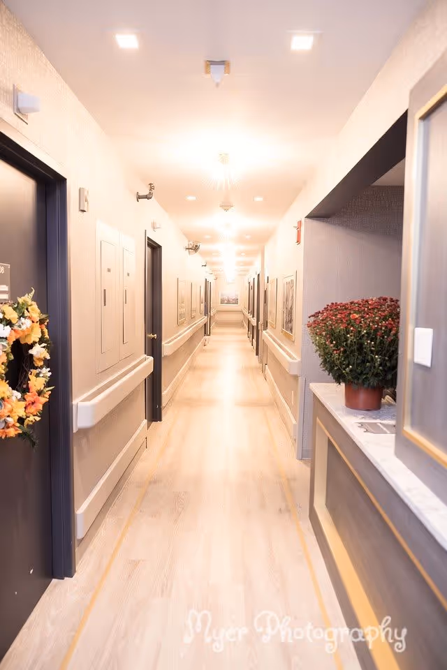 Bright, long indoor hallway in a senior living facility with doors, handrails, and a reception counter with a potted plant.