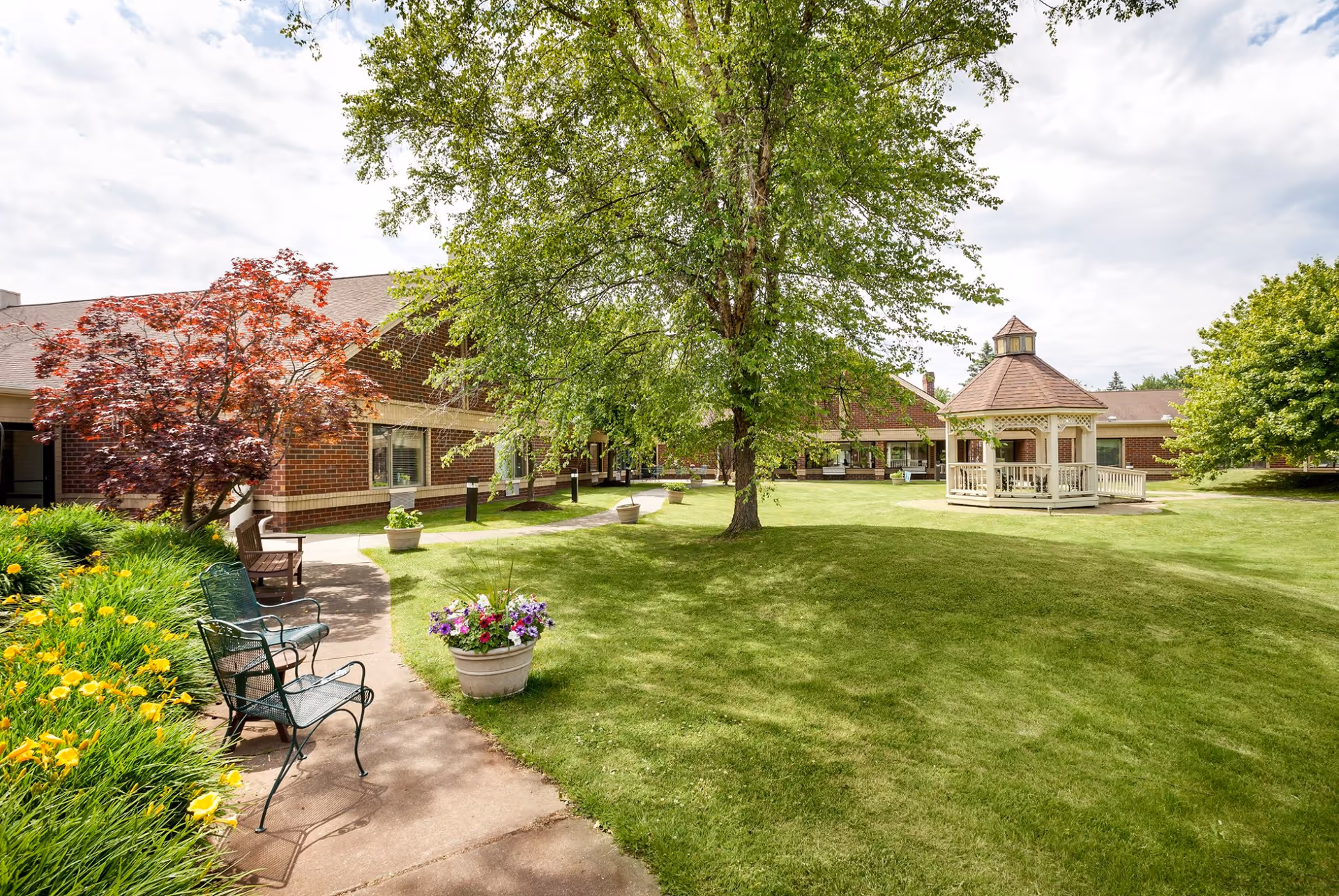 A well-maintained outdoor garden area at Pomeroy Living Sterling featuring a large green lawn, a central tree, a white gazebo, flower pots with colorful flowers, benches, and a brick building in the background under a partly cloudy sky.