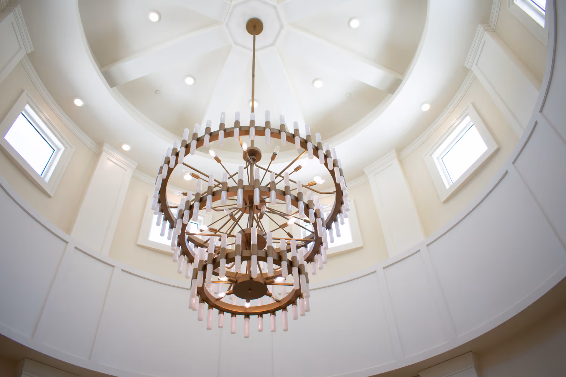A large, ornate chandelier with multiple circular tiers and numerous small light bulbs hangs from a high, white, domed ceiling with recessed lighting and several small windows around the perimeter.