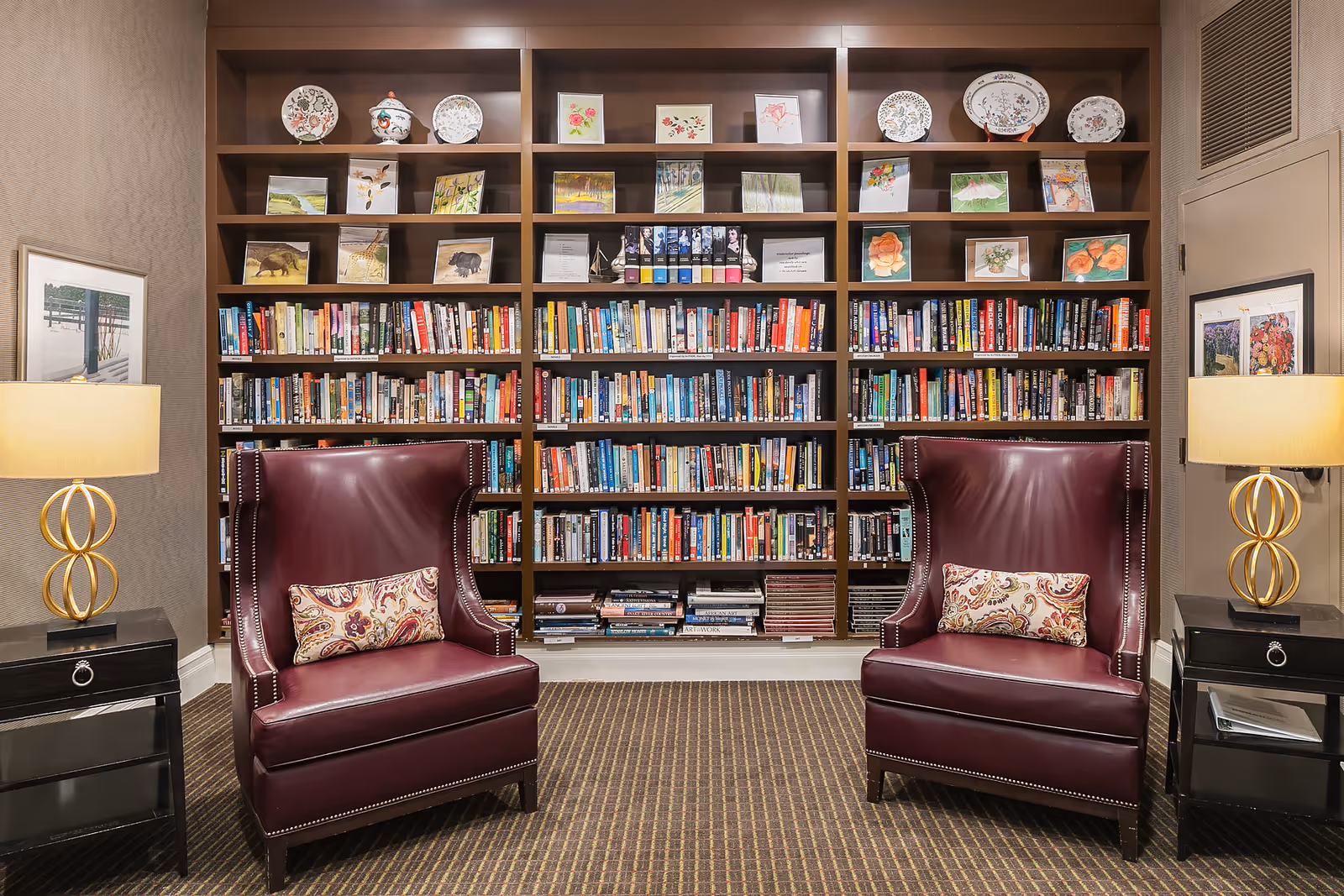 A cozy reading area with two burgundy leather armchairs, each with a decorative pillow, positioned in front of a large wooden bookshelf filled with books and decorative plates and artwork on the top shelves. Two side tables with modern lamps flank the chairs, and framed pictures hang on the walls.