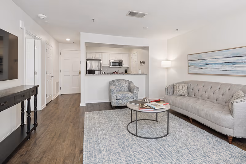 A modern living room with a gray tufted sofa, a patterned armchair, a round coffee table with books and decorative items, a floor lamp, and a large abstract painting on the wall. The room has wood flooring and a light blue area rug. In the background, there is an open kitchen with white cabinets, stainless steel appliances, and a granite countertop.